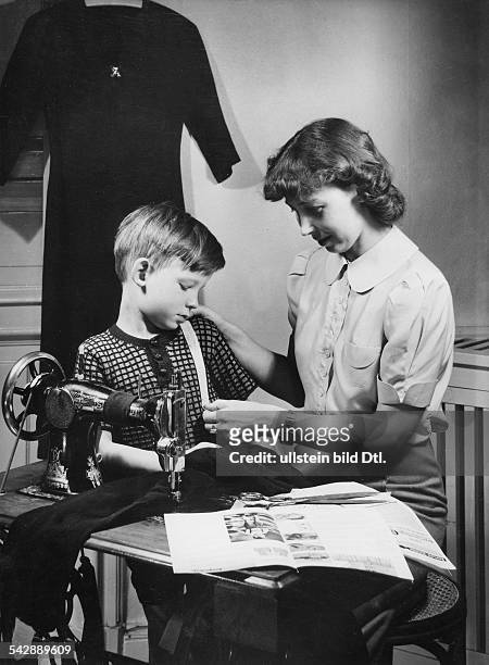 Frau misst den Oberkörper ihres Sohnes ab, um ein Kleidungsstück dementsprechend an der Nähmaschine zu ändern1939