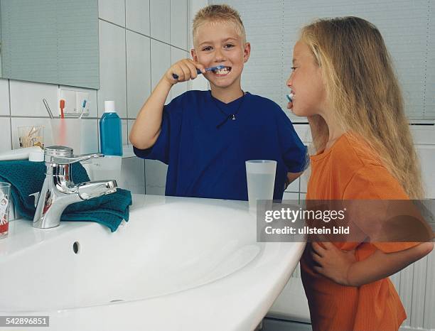 Zwei Kinder, Junge und Mädchen, stehen gemeinsam am Waschbecken im Badezimmer und reinigen ihre Zähne. Auf dem Waschbeckenrand stehen mit Wasser...