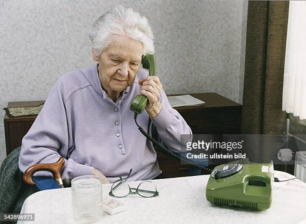 Eine Seniorin am Telefon. Vor ihr auf dem Tisch steht ein Trinkglas und eine Aufbewahrungsbox für Tabletten; davor liegt eine Brille. Am Stuhl der...