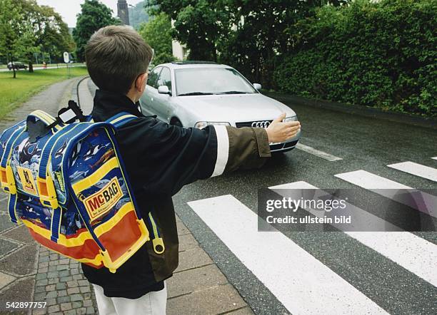 Mit erhobener Hand steht ein Kind an einem Zebrastreifen. Ein PKW stoppt, damit der Junge die Straße überqueren kann. Der junge Verkehrsteilnehmer...