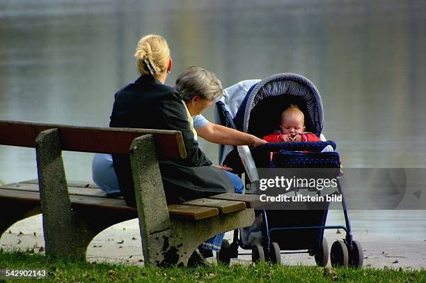 Mit dem Kind im Kinderwagen ruhen sich die Frauen auf einer Bank am See aus. Generationen; Baby; Kleinkind; Oma.