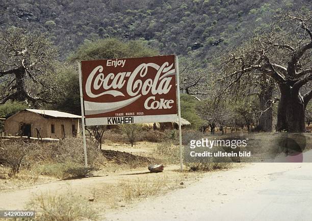 Am Ortsausgang eines tansanischen Dorfes steht ein großes Werbeschild mit der Aufschrift "Enjoy Coca-Cola Coke". Darunter befindet sich in Kiswahili...