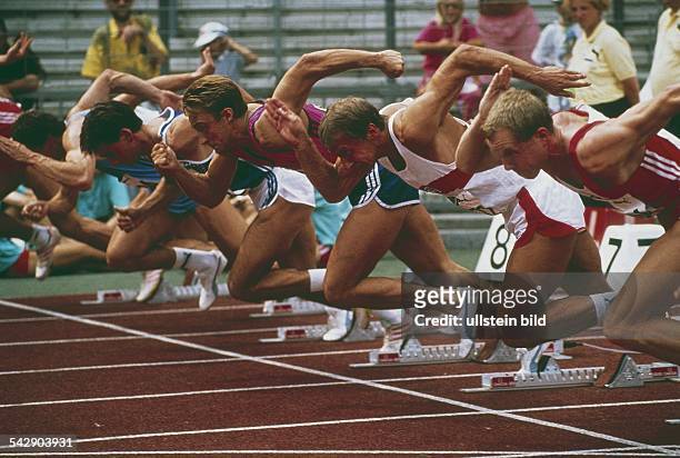 Eine Gruppe Läufer prescht kraftvoll nach dem Startschuss aus den Startblöcken. Ihre Rücken sind gebeugt, die muskulösen Arme werden in die...
