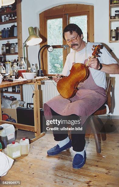 Instrumentenbauer Hubert Schnorr, bekleidet mit einer Schürze, sitzt in seiner Werkstatt und lackiert mit einem Pinsel eine Geige, die er auf dem...