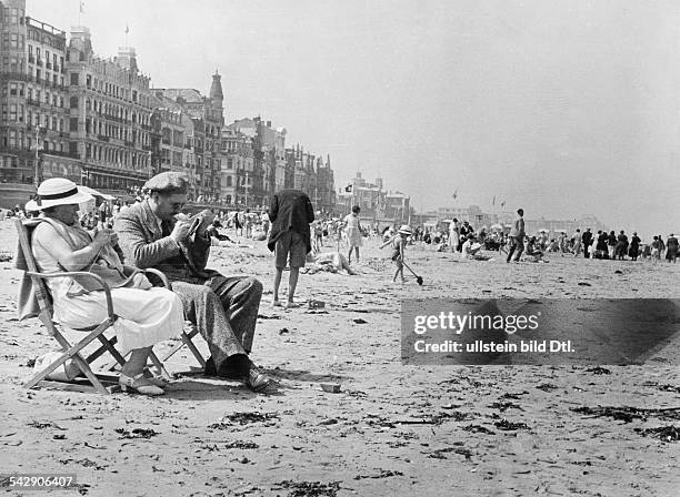 Strandleben in Ostende: ein Paar auf Klappstühlen; die Frau strickt, der Mann löst ein Kreuzworträtsel in einer Zeitungveröffentlicht: Dame,...