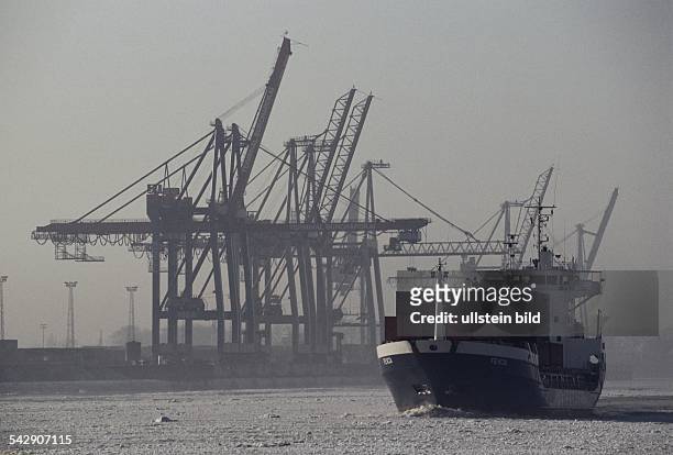 Hamburg: ein Containerschiff fährt auf der vereisten Elbe. Links die Ladebäume und Kräne des Burchardkais. Es liegt leichter Nebel über dem Fluss. .