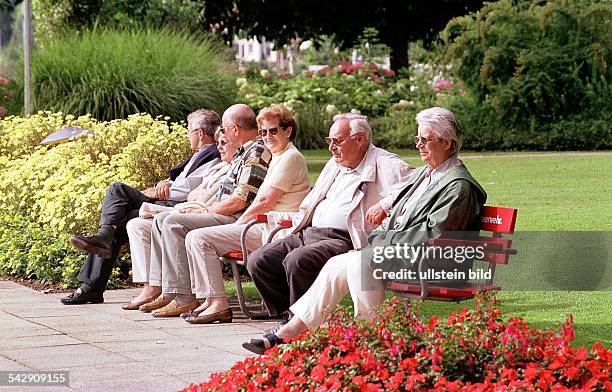 Eine Gruppe alter Leute genießt nebeneinander auf einer Parkbank sitzend die Sonne und die Idylle der sie umgebenden Blumenpracht. Rentner; Senioren....