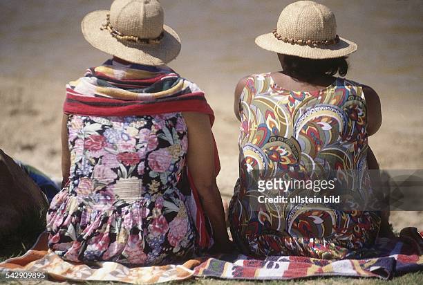 Rückansicht von zwei Frauen in der südafrikanischen Kap Provinz. Sie tragen bunte Kleider sowie Strohhüte und sitzen auf einem Handtuch auf dem Boden.