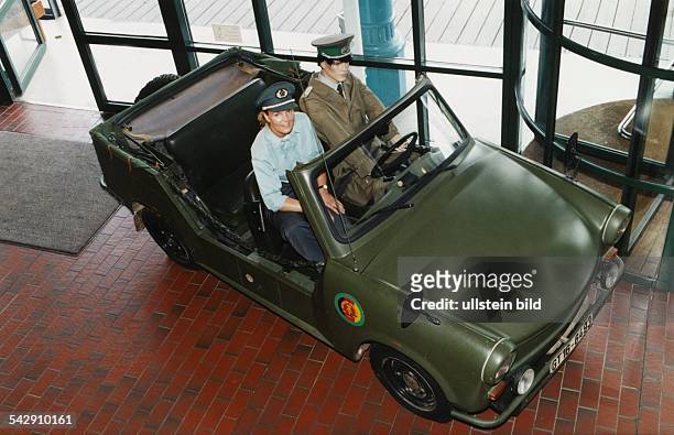 Der Blick von oben auf einen olivgrünen Trabant Cabrio, in dem eine Puppe sitzt, die einen DDR-Zollbeamten in Uniform mit Mütze darstellt. Daneben...