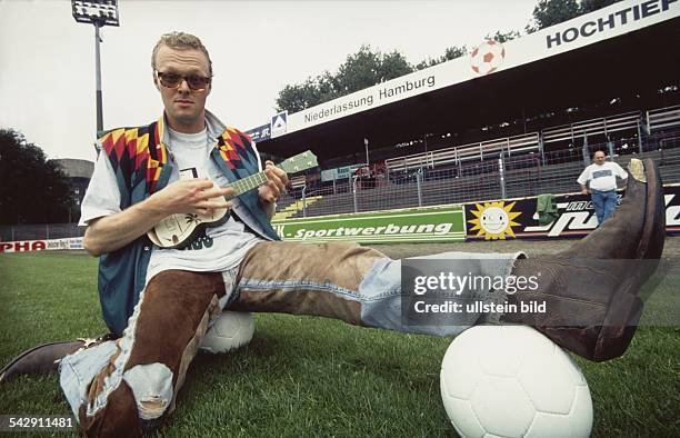 Der Moderator und Sänger Stefan Raab mit Ukulele, auf zwei Fußbällen sitzend, im Fußballstadion. Aufgenommen um 1994.