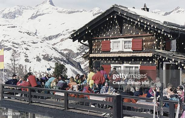 Eine Berghütte aus Holz mit roten Fensterläden. Skiläufer und Skitouristen sitzen auf der Terrasse und genießen die Sonne. Im Hintergrund ein...