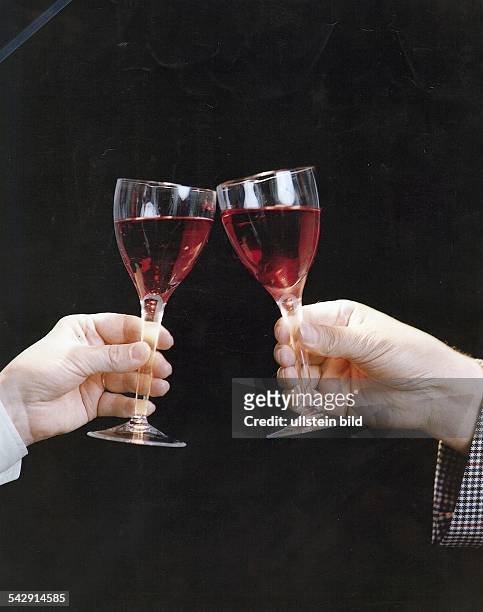 Zwei Hände mit Weingläsern, mit denen angestossen wird. In den Gläsern befindet sich Rotwein. Undatiertes Foto.