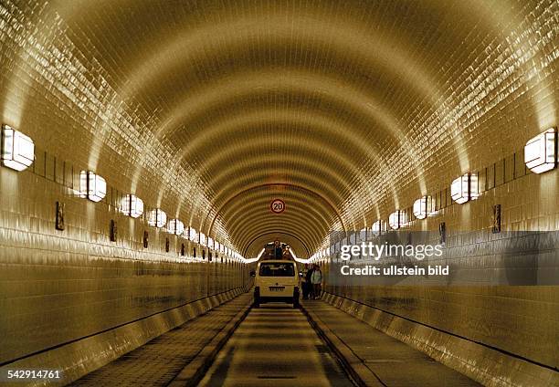 Hamburg: eine der weißgefliesten Röhren des Alten Elbtunnels. Links und rechts die Art Deco-Beleuchtung. Auf der engen Fahrbahn fährt ein Auto, auf...