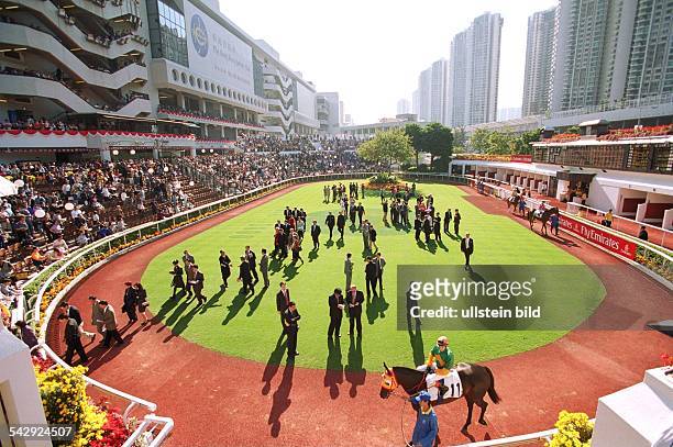 Galopprennbahn "Sha Tin" in Hongkong, umgeben von Hochhäusern. Pferde mit Jockeys auf der roten Sandbahn werden von Zuschauern auf der Rasenfläche...