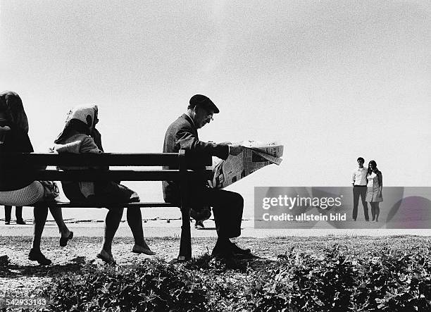 älterer Mann sitzt zusammen mit Frauen auf einer Bank am Strand und liest in einer Zeitung, im Hintergrund steht ein junges Paar- undatiert, um 1970