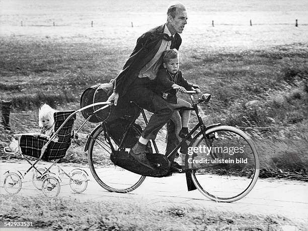 Ein Vater mit seinem Sohn auf dem Fahrrad; gleichzeitig zieht er den Kinderwagen mit seiner Tochter hinter sich her- 1973