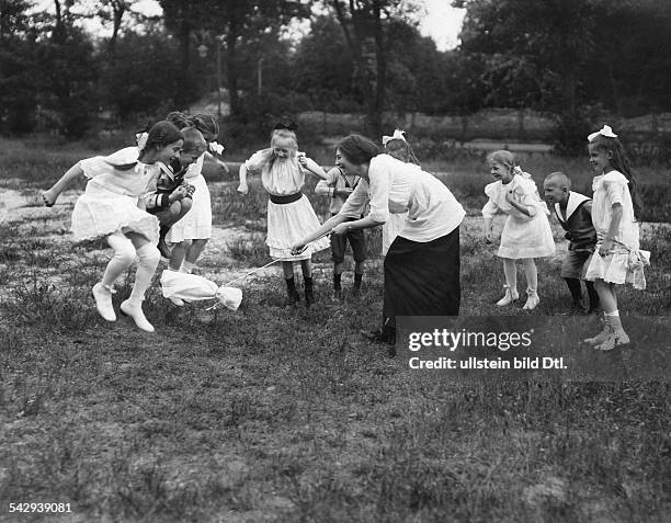 Hüpfspiel: Kinder müssen über den Beutel springen, den eine Frau in der Mitte kreisen lässt.- um 1915Foto: Hünich, Conrad