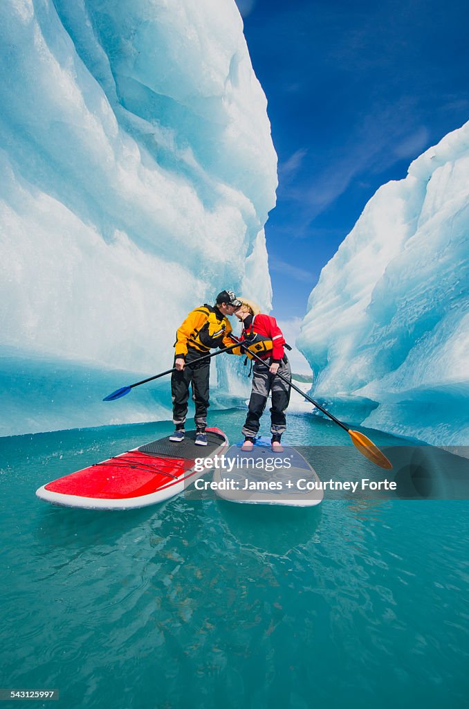 A couple on stand up paddle boards (SUP) kiss in an iceberg canyon on Bear Lake in Kenai Fjords National Park, Alaska.