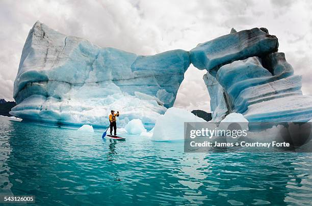 one man on stand up paddle board (sup) paddles past hole melted in iceberg on bear lake in kenai fjords national park, alaska. - los glaciares national park - fotografias e filmes do acervo