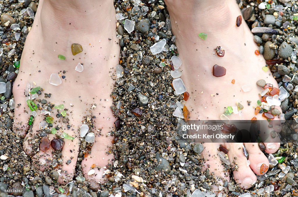 A mans feet are covered in glass from the handfuls of beach glass that wash ashore at Glass Beach in Fort Bragg, California.