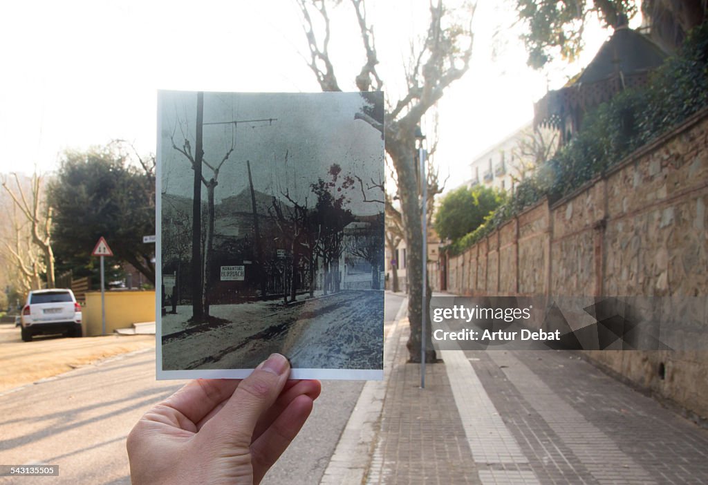 Now and then, holding picture by hand on street.