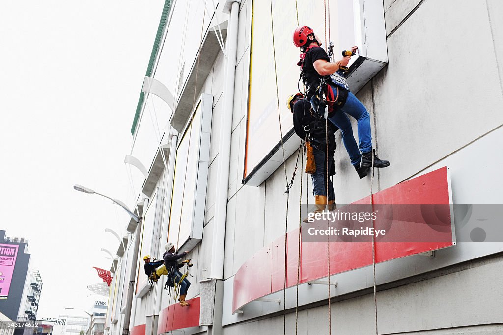 Workers rappelling down a shopping mall exterior to access billboards