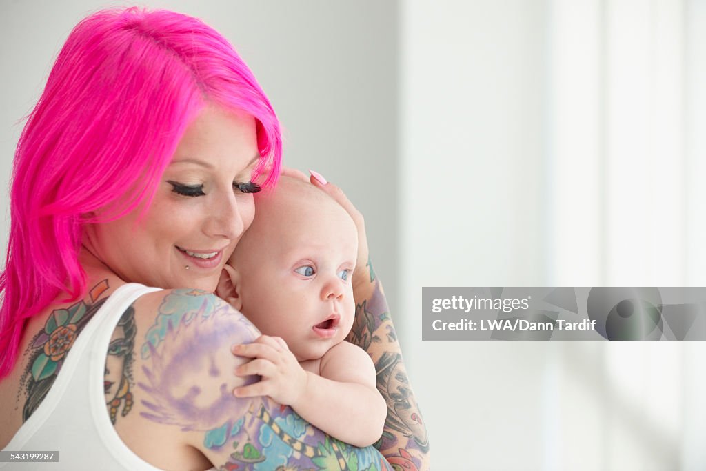 Caucasian mother with pink hair and tattoos cradling baby