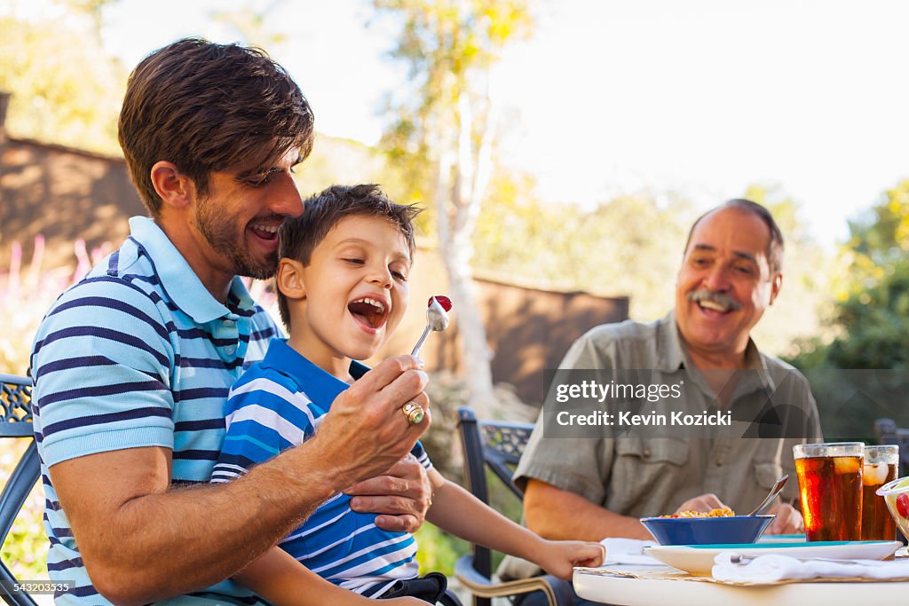 Father feeding son on lap, grandfather in background