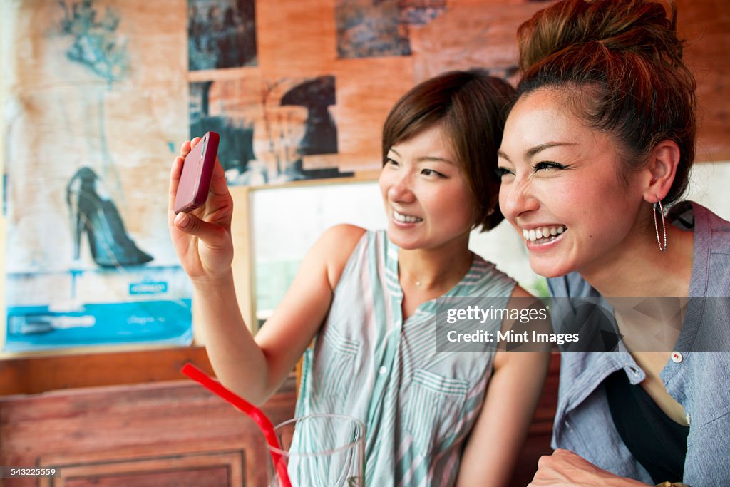 Two women looking at a cell phone, taking a selfie, sitting indoors. 