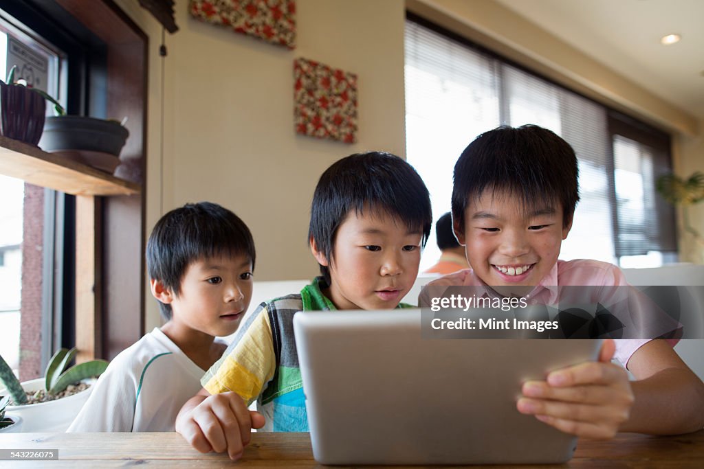 Three boys sitting at a table, looking at a digital tablet, smiling.