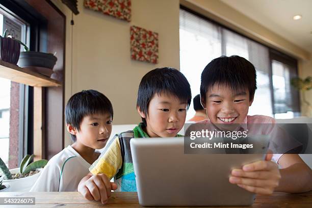 three boys sitting at a table, looking at a digital tablet, smiling. - 子供のみ ストックフォトと画像