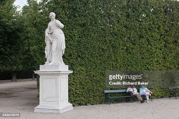 Ladies sit to rest on a bench with sore feet beneath a statue at Schloss Schonbrunn on 27th June 2016, in Vienna, Austria. Schonbrunn is a former...