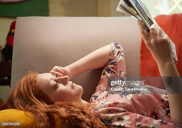 young woman lying on sofa at home reading magazine - woman reading magazine stock pictures, royalty-free photos & images