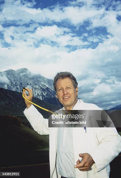 Der "Bergdoktor" von SAT 1, Gerhard Lippert, steht im weißen Ärztekittel vor dem Großfoto eines Bergmassivs. Er hält das Stethoskop an die Abbildung...