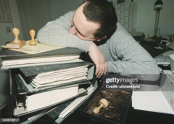Ein Beamter schläft an seinem Schreibtisch auf einem Stapel Aktenordnern. Neben ihm liegen Stempel und eine Schachtel mit Büroklammern. Aufgenommen...