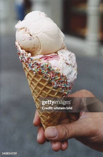 Eine Eiswaffel mit zwei Kugeln Milchspeiseeis, gehalten von einer Hand. Der Rand der Eiswaffel ist verziert mit bunten Streußeln. .