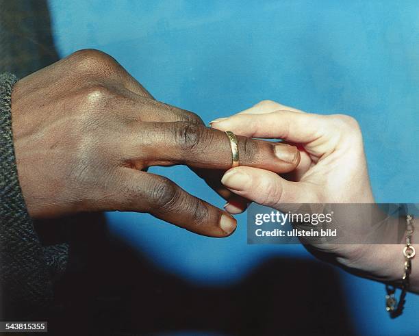 Eine weiße Frauenhand steckt einen Ehering auf den Ringfinger einer schwarzen Männerhand. Scheinehe; Ehe mit einem Ausländer; Mischehe. Symbolbild .