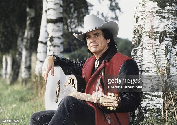 Der Musiker Tom Astor sitzt mit einer Gitarre auf dem Schoß an einem Birkenstamm gelehnt. Er trägt einen Cowboyhut und für Countrymusik typische...