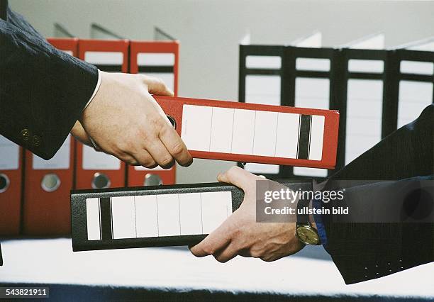 In den Händen zweier Männer befinden sich ein roter und ein schwarzer Aktenordner, die getauscht werden. Im Hintergrund stehen ebenfalls Aktenordner....