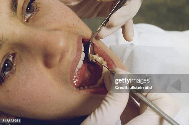 Eine Patientin sitzt mit weit geöffnetem Mund beim Zahnarzt. Mit Hilfe eines Mundspiegels und eines Zahnreinigungsinstrumentes werden ihre Zähne auf...