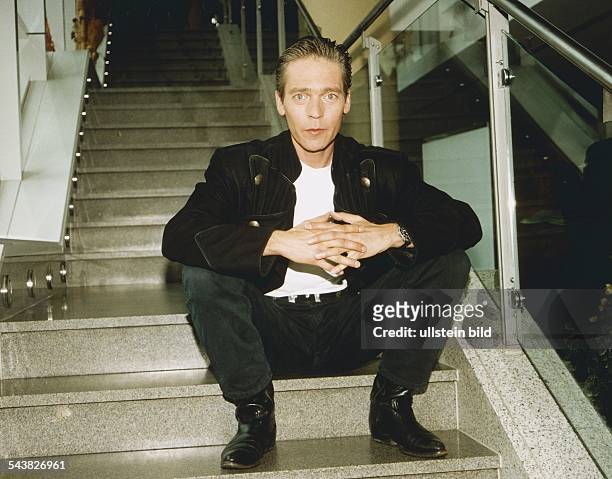 Der deutsche TV- und Bühnenschauspieler aus München sitzt mit gefalteten Händen auf einer Treppe. Er trägt eine bayrische Trachtenjacke und Stiefel....