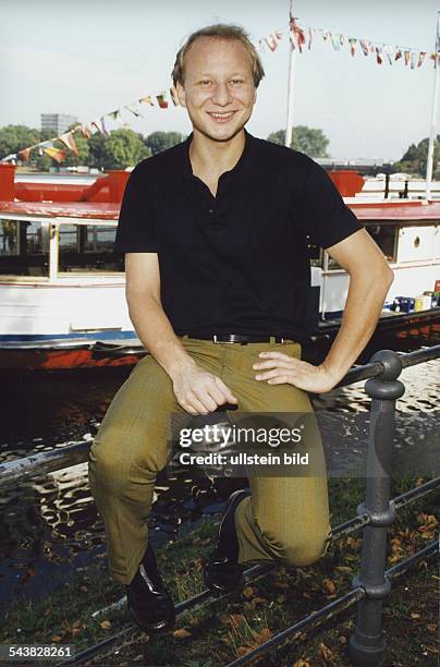 Schauspieler Christopher Zumbült sitzt auf einem Metallzaun an der Binnenalster. Er trägt ein schwarzes Polohemd und eine karierte Hose. Im...