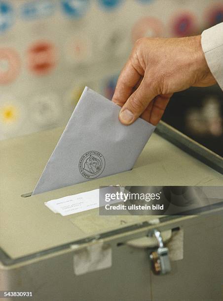 Eine Hand steckt einen Umschlag in eine Wahlurne. Der Briefumschlag trägt einen Stempel mit der Aufschrift "Land Nordrhein-Westfalen". Die Wahlurne...