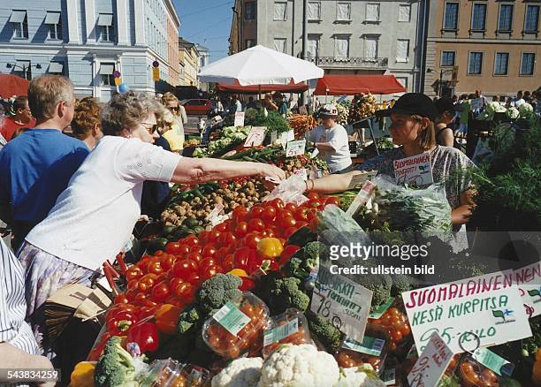Eine Kundin beugt sich über die reiche Auslage eines Gemüsemarktes in Helsinki um ihre Ware zu bezahlen. Das Angebot besteht unter anderem aus...