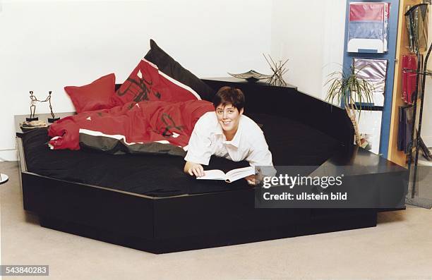 Eine Frau liegt auf einem sechseckigen, schwarzen Wasserbett. Auf dem Bett liegt rotschwarze Bettwäsche in japanischem Design. .