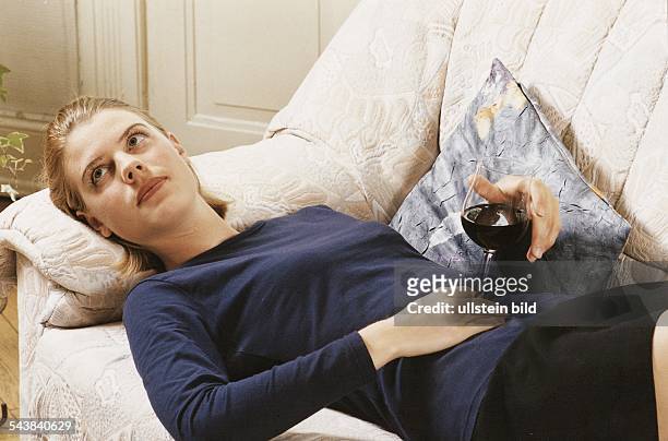 Eine junge Frau liegt in legerer Kleidung und mit traurigem Gesichtsausdruck auf einem Sofa. In der Hand hält sie ein Glas Rotwein. Traurigkeit;...