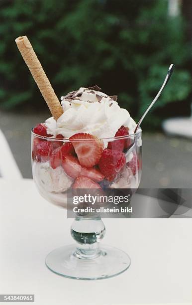 Erdbeereis mit frischen Erdbeerstückchen in einem Glas, dekoriert mit Sahne, Schokoladenstreußeln und einem Waffelröllchen. Rechts steckt ein Löffel....