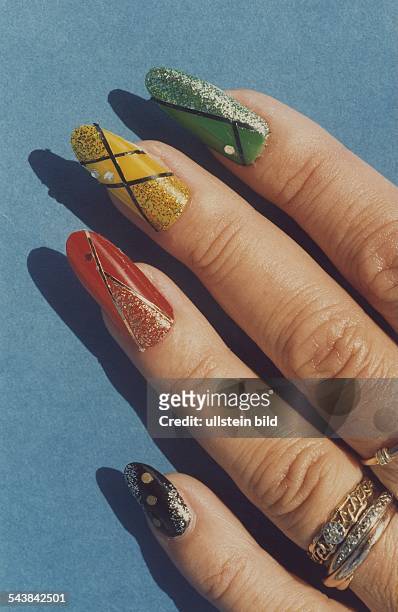 Eine Frauenhand mit langen, lackierten Fingernägeln unterschiedlicher Farbe. Auf die Nägel sind Straß, Glitter und Silberfäden geklebt. Entwurf: Rita...