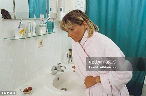 Badezimmer: eine junge Frau, mit Bademantel bekleidet, lehnt sich an ein Waschbecken und hält sich den Magen. Übelkeit; Magenschmerzen;...