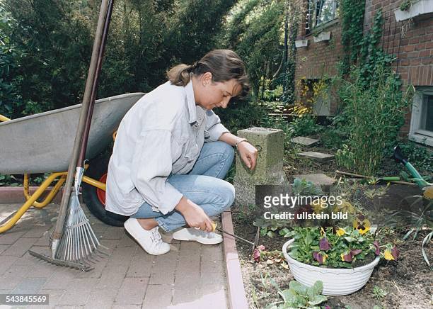 Eine junge Frau jätet im Garten Unkraut mit einer Kombihacke. Neben ihr steht eine Schubkarre, an die eine Harke und ein Rechen angelehnt sind....